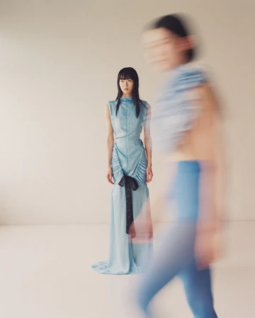 Two young Asian women pose in a light studio setting. One stands in focus wearing a blue dress with a black bow. Soft colors.