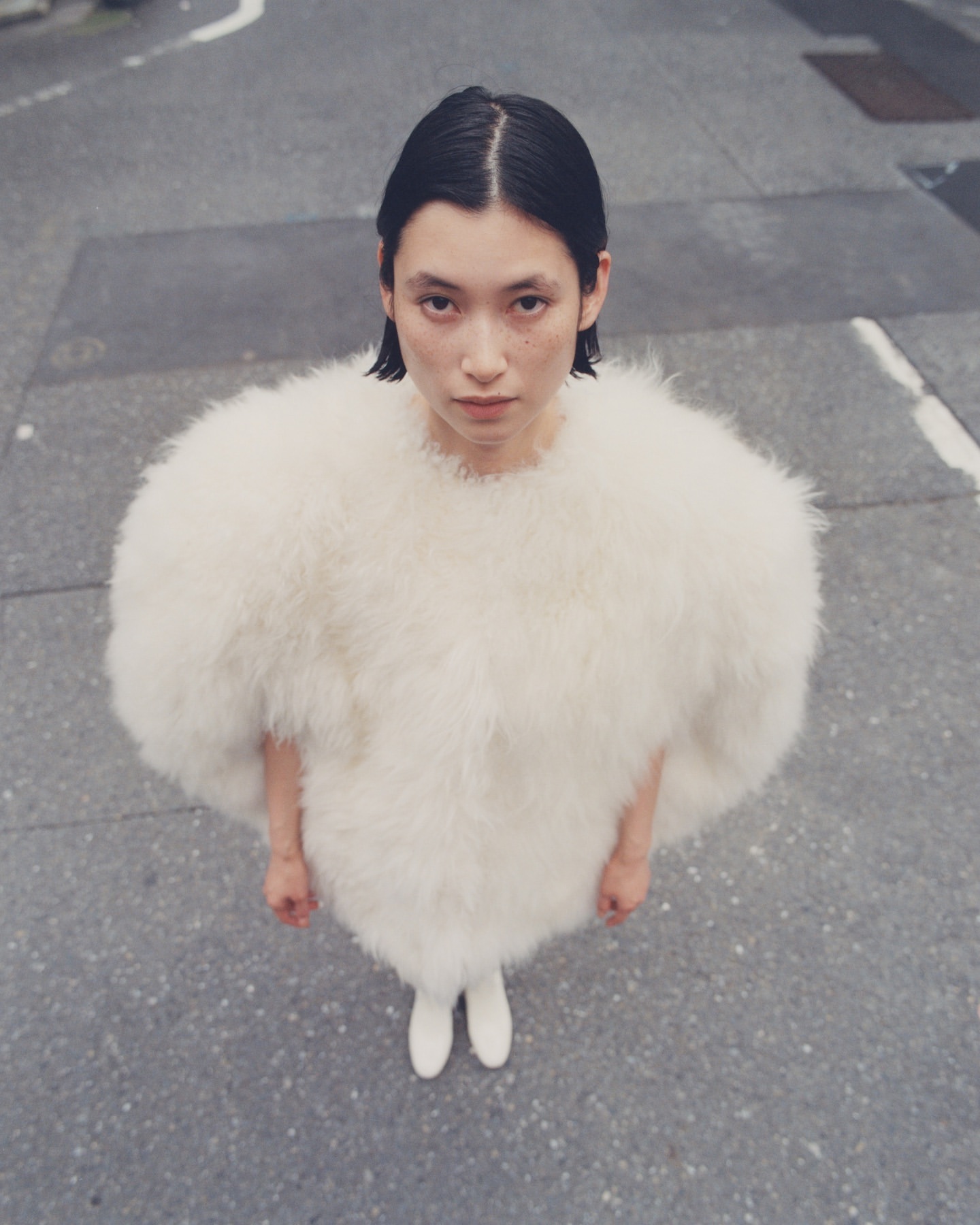 An Asian adult female stands outdoors on a city street, wearing a puffy white feather jacket and white shoes, shot from a high angle.