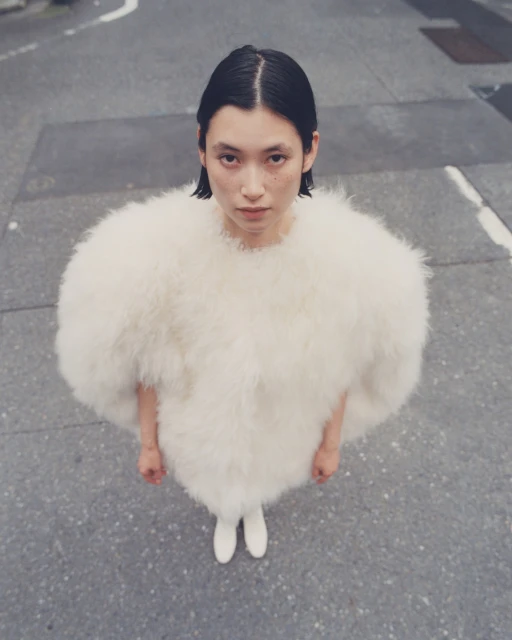 An Asian adult female stands outdoors on a city street, wearing a puffy white feather jacket and white shoes, shot from a high angle.