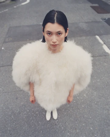 An Asian adult female stands outdoors on a city street, wearing a puffy white feather jacket and white shoes, shot from a high angle.