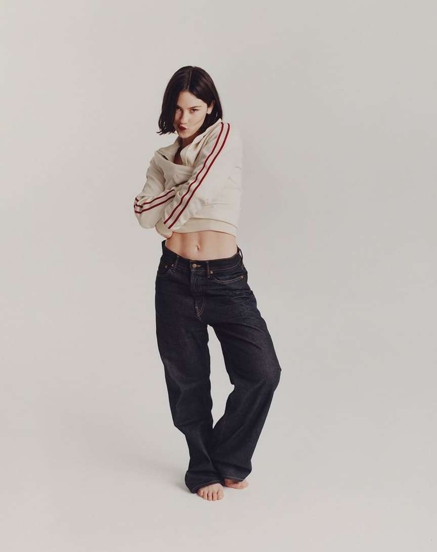 Woman posing in a light sweatshirt with red stripes and baggy dark jeans in a white studio with arms crossed, looking at the camera.