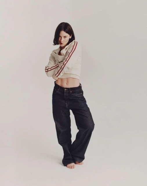 Woman posing in a light sweatshirt with red stripes and baggy dark jeans in a white studio with arms crossed, looking at the camera.