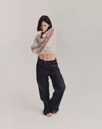 Woman posing in a light sweatshirt with red stripes and baggy dark jeans in a white studio with arms crossed, looking at the camera.