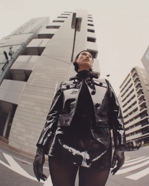Woman in glossy black jacket and gloves poses in an urban fashion shot with tall buildings at eye-level view in the daytime.