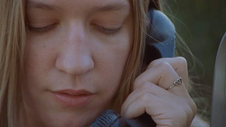 Close-up shot of a woman with fair skin, fair hair, and closed eyes talking on a black landline phone with a silver heart ring on her finger.