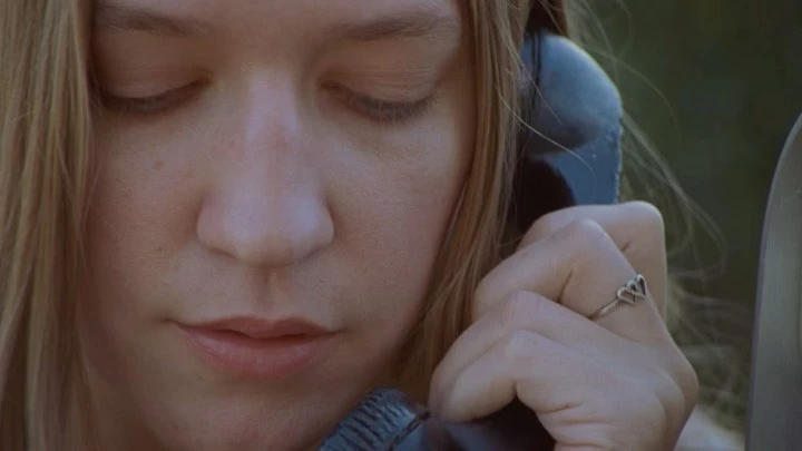 Close-up shot of a woman with fair skin, fair hair, and closed eyes talking on a black landline phone with a silver heart ring on her finger.