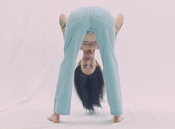 Woman bends forward in blue jeans against a bright white backdrop. She has dark hair, fair skin, and tattoos on her arms.