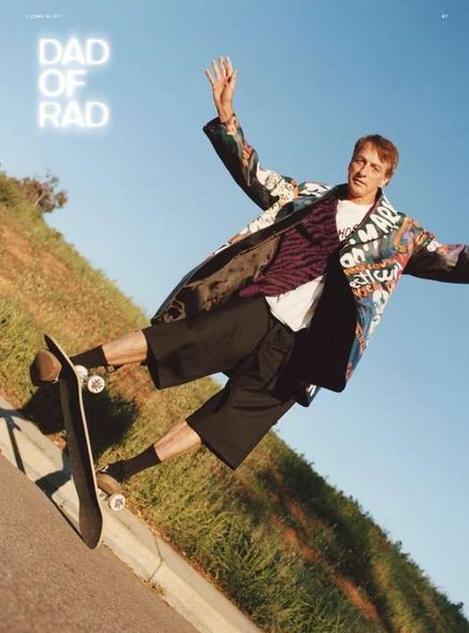 Tony Hawk executes a skateboarding trick with arms outstretched against a blue sky, captured at a low angle with a grassy hill backdrop.