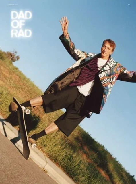 Tony Hawk executes a skateboarding trick with arms outstretched against a blue sky, captured at a low angle with a grassy hill backdrop.