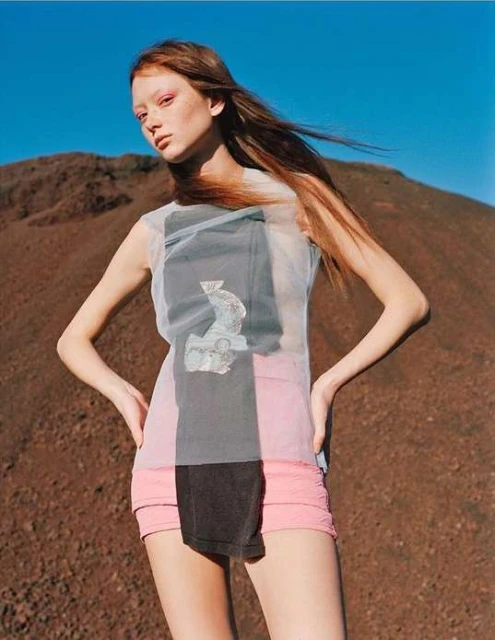 A young, thin fashion model poses in a sleeveless top and short skirt against a brown dirt pile under a clear blue sky in an outdoor setting.