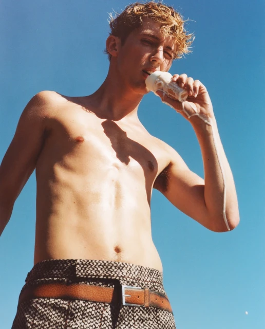 Low-angle shot of a shirtless blond man eating ice cream against a bright blue sky, wearing patterned pants with a brown belt.