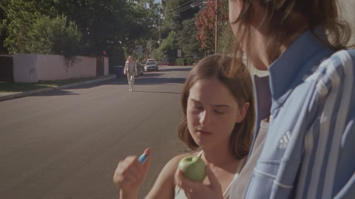 Two young adults on a tree-lined street, one holding a green apple and blue capsule, while another person walks in the background.