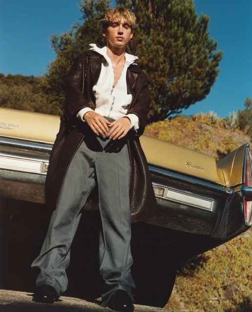 Young adult male poses with a vintage yellow Cadillac wearing a long brown leather coat, white zip-up hoodie and gray pants outdoors.