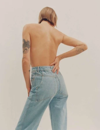 Model with tattoos on back and arms wearing light blue denim jeans against a plain white studio backdrop.