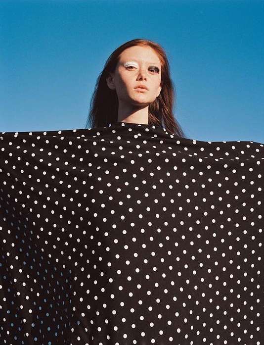 Adult female with red hair, wearing black-and-white polka dot fabric against bright blue sky, posing with dramatic makeup in a studio shot.