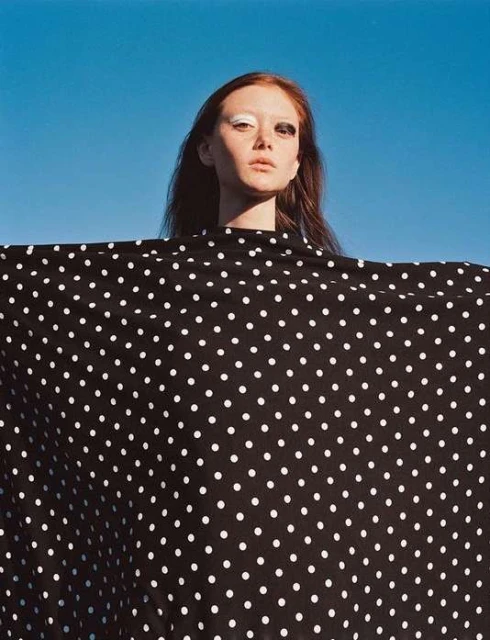 Adult female with red hair, wearing black-and-white polka dot fabric against bright blue sky, posing with dramatic makeup in a studio shot.