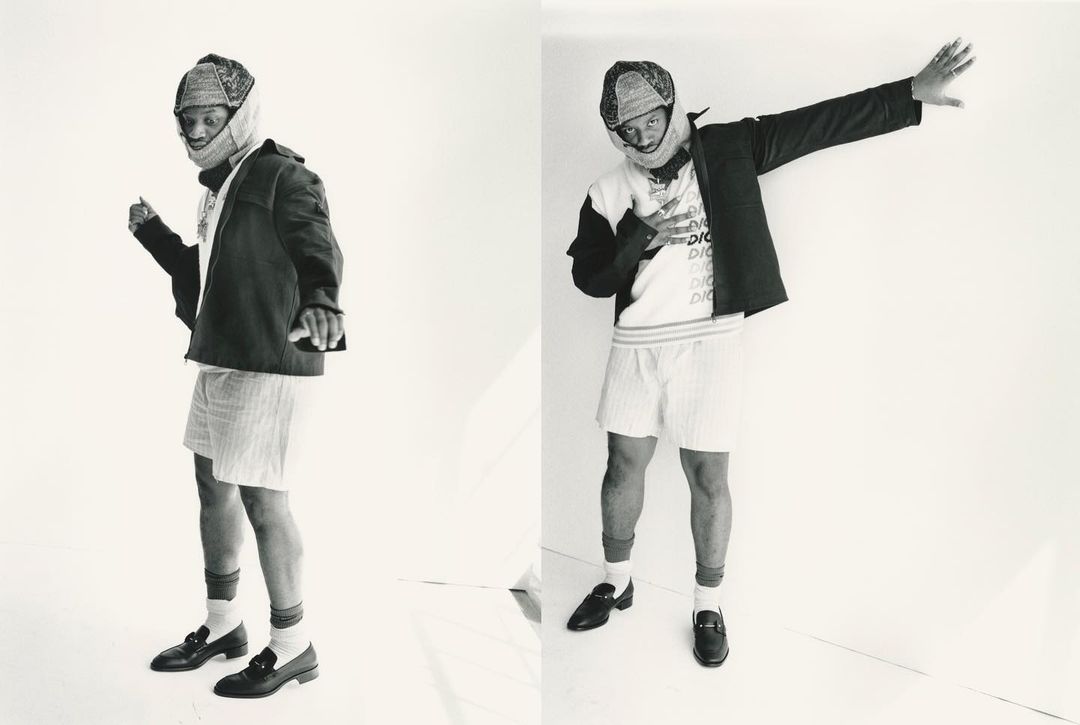 A Black male fashion model poses in a jacket, shorts, and loafers, in a photo studio setting, with a white backdrop. Fashion editorial.