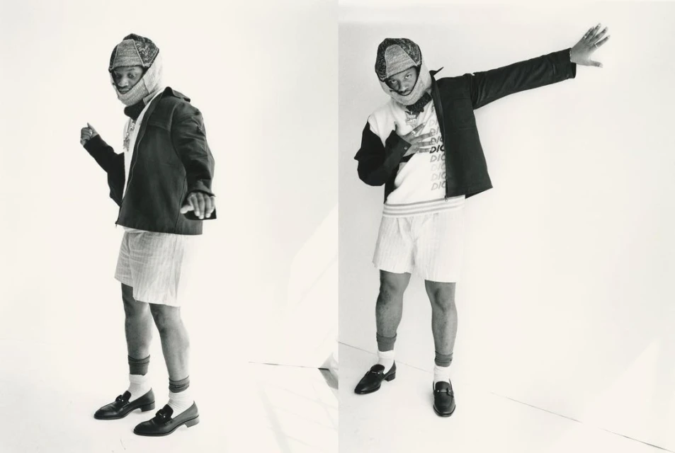 A Black male fashion model poses in a jacket, shorts, and loafers, in a photo studio setting, with a white backdrop. Fashion editorial.