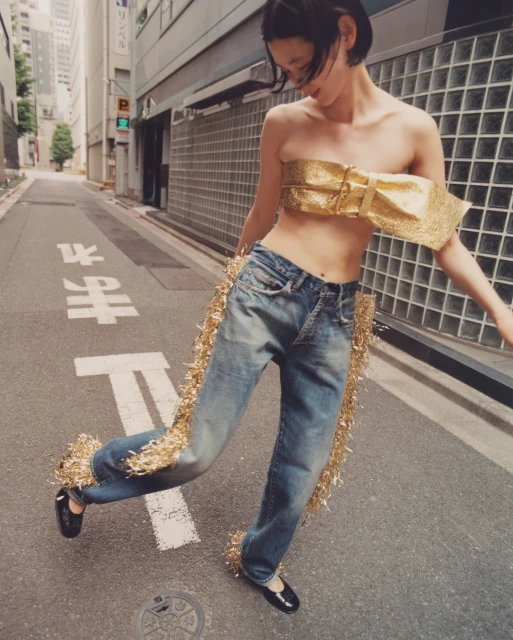 A woman with a short haircut dances in a city street. She wears a gold top, distressed blue jeans with gold tinsel, and black flats.