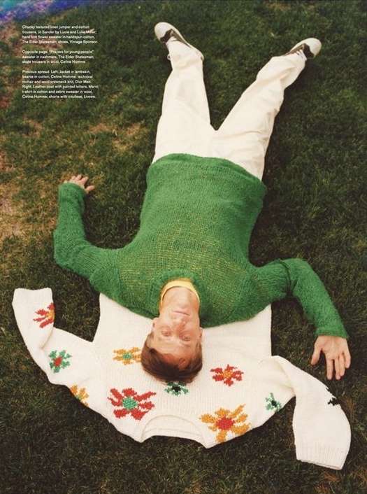 An adult male is lying in green grass wearing a green sweater, with a cream-colored sweater with flowers laying next to him.