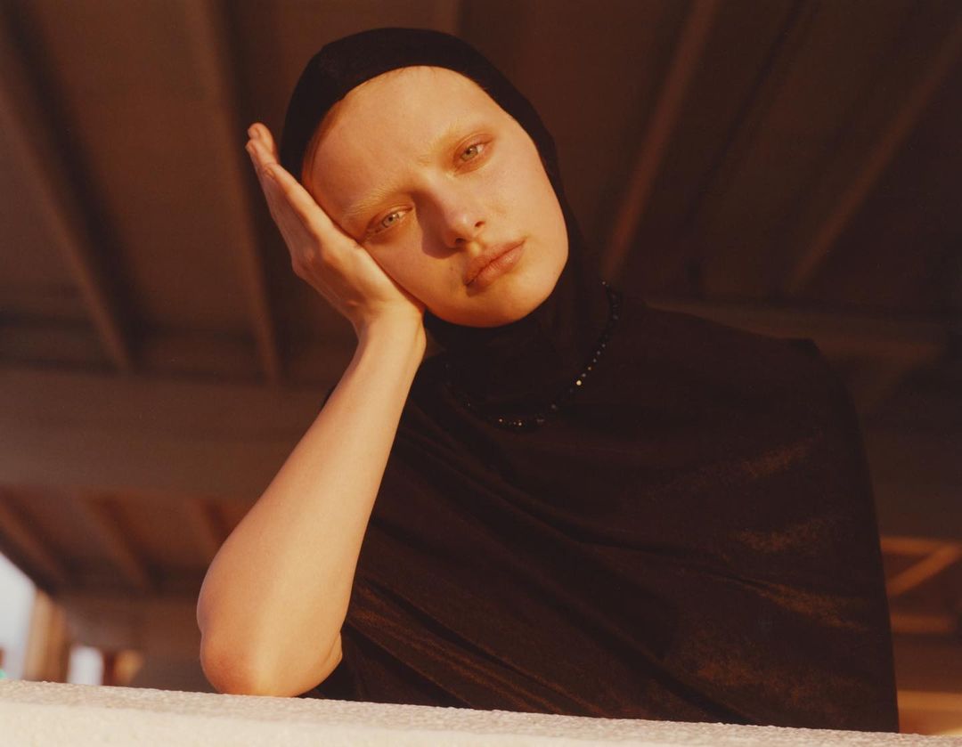 Close-up portrait of an adult woman with fair skin, black headscarf, and matching turtleneck, resting her face on her hand.
