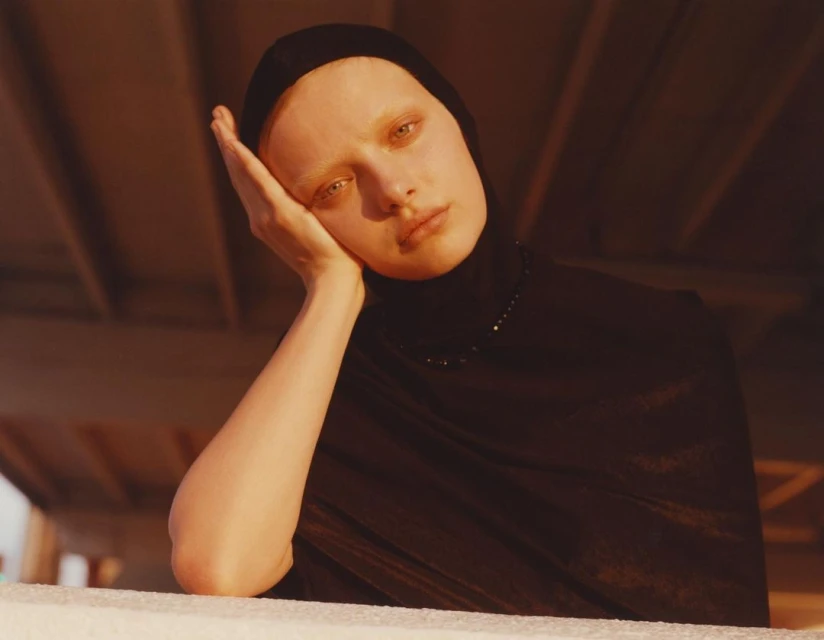 Close-up portrait of an adult woman with fair skin, black headscarf, and matching turtleneck, resting her face on her hand.