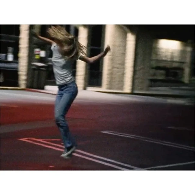 An adult female jumps in the air in an outdoor parking lot at night, with a blurred building and light providing illumination.