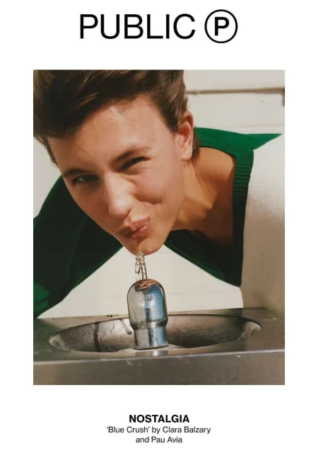 Close-up of a young adult female drinking from a metal public water fountain outdoors, wearing a white and green sweater, front view.