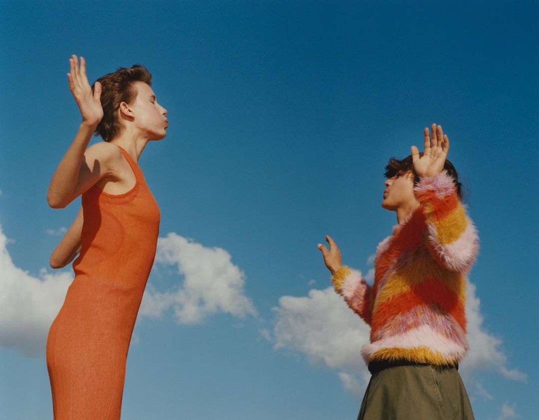 Two women stand outdoors against a blue sky with clouds; one wears an orange dress, the other a colorful striped sweater.
