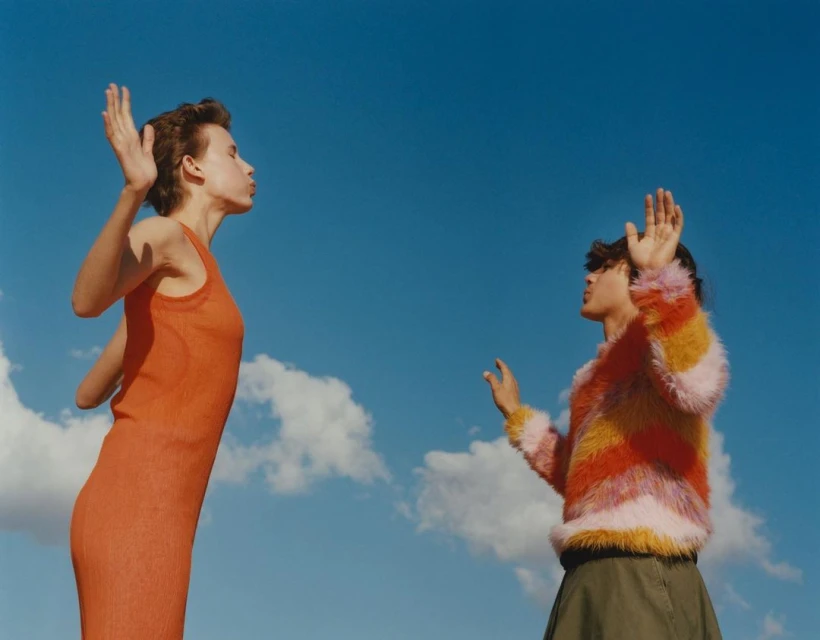Two women stand outdoors against a blue sky with clouds; one wears an orange dress, the other a colorful striped sweater.