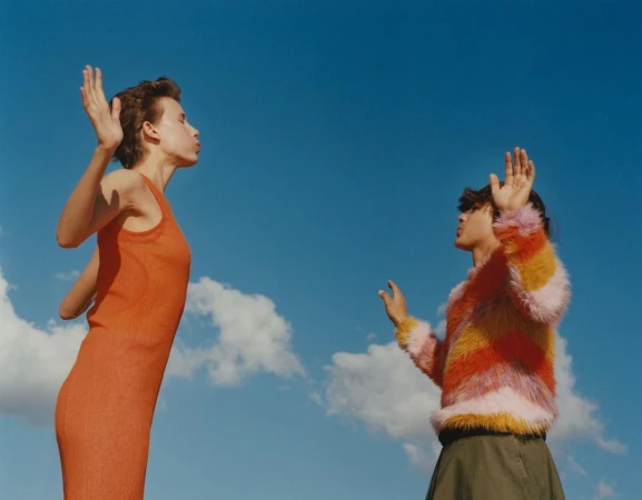 Two women stand outdoors against a blue sky with clouds; one wears an orange dress, the other a colorful striped sweater.