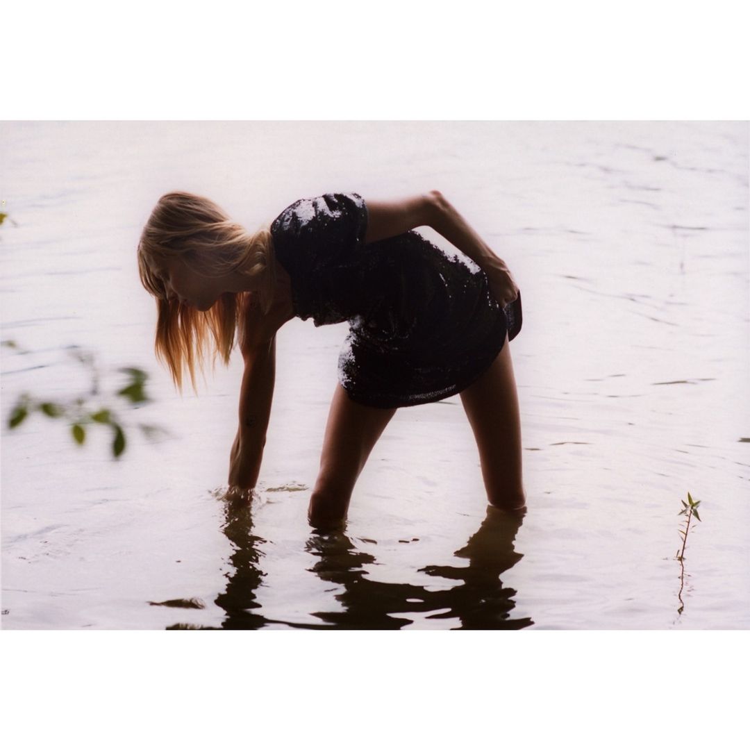 Woman in a black dress stands in shallow water, bending over with her hand reaching into the water, captured in a medium shot.