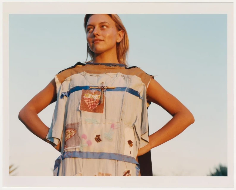 Young woman in handmade patchwork dress poses outdoors against a clear sky, showcasing fashion and creative design.
