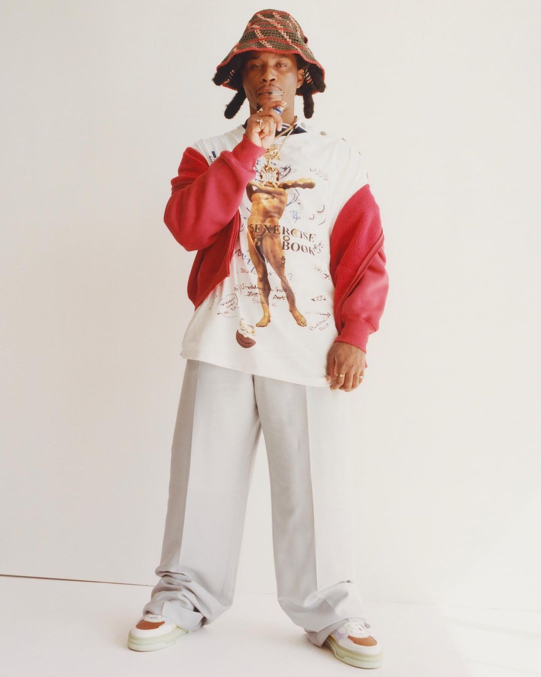 Full body shot of a Black adult male in a colorful bucket hat, patterned shirt, gray pants, and sneakers against a white backdrop.