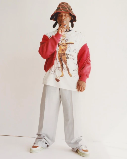 Full body shot of a Black adult male in a colorful bucket hat, patterned shirt, gray pants, and sneakers against a white backdrop.