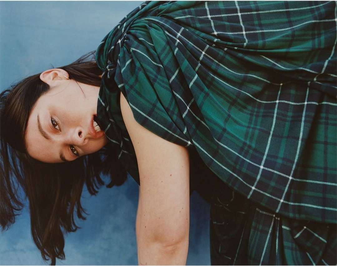 A woman with dark hair is draped in a green and navy plaid fabric against a blue background, creating a fashion-forward and visually striking image.