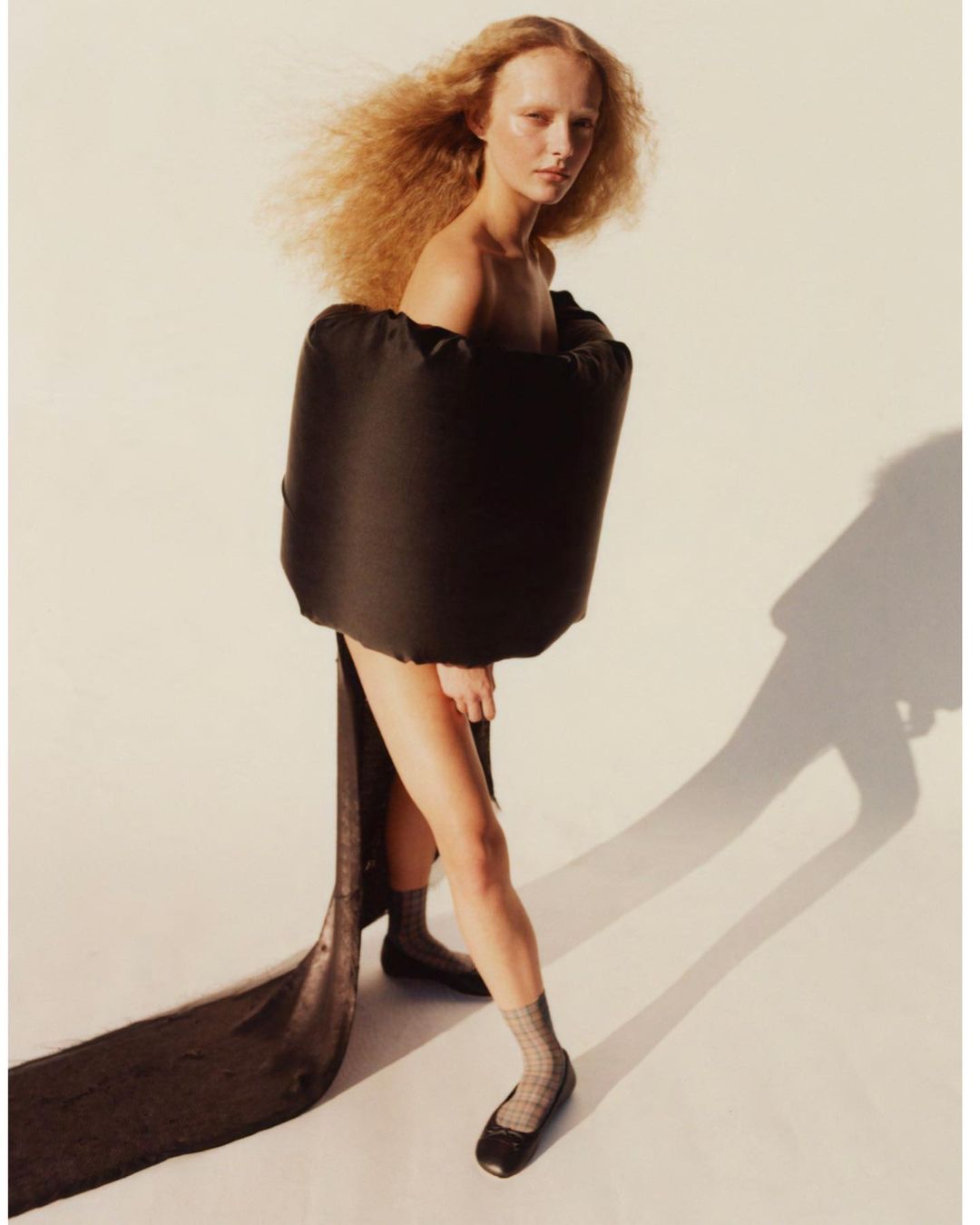 Fashion shot of a woman with curly hair wearing a black dress and flats in a studio, capturing a blend of modern style and elegance.