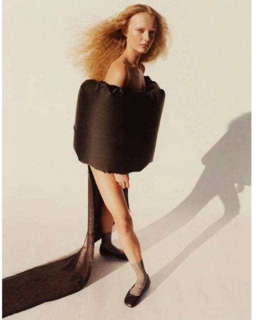 Fashion shot of a woman with curly hair wearing a black dress and flats in a studio, capturing a blend of modern style and elegance.
