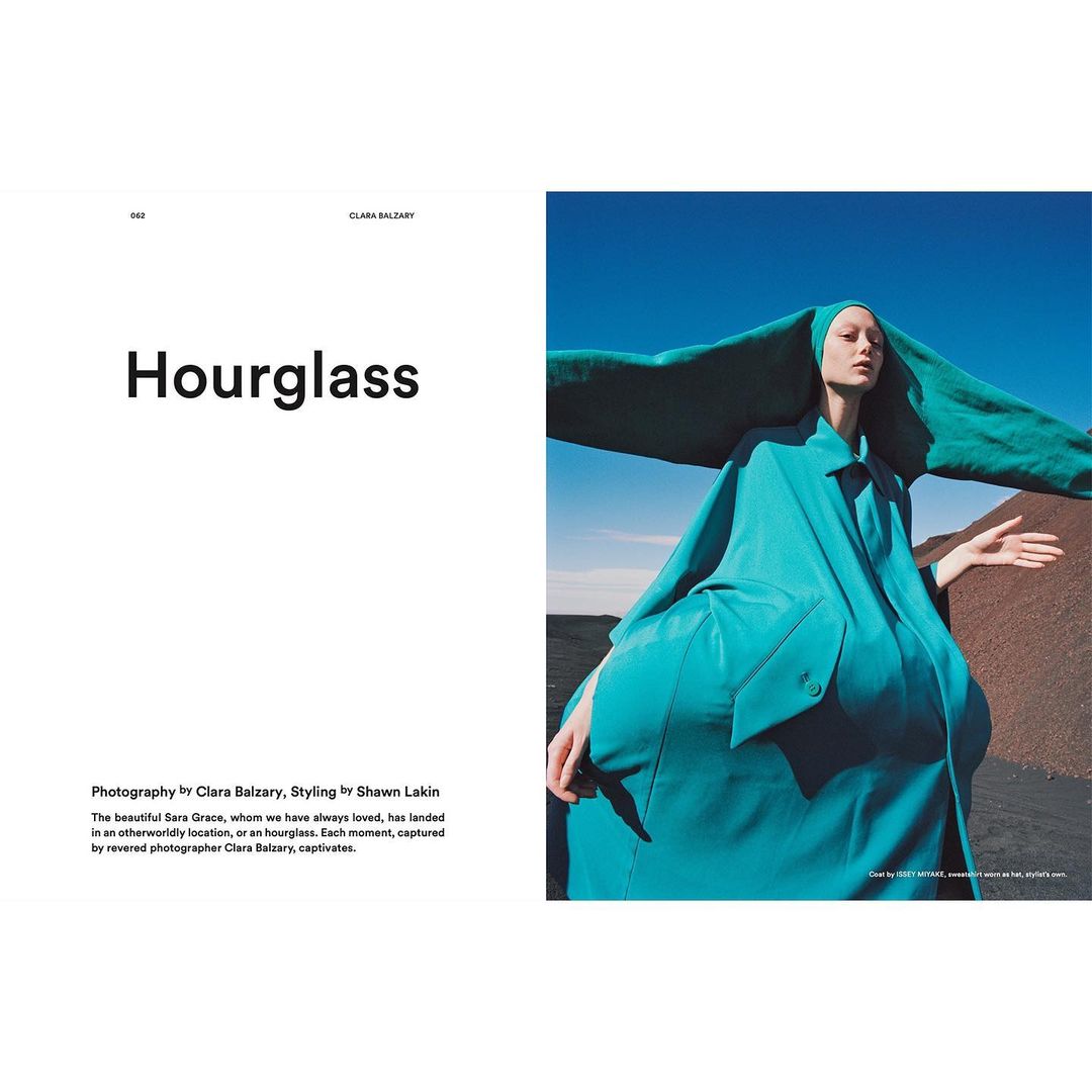 Fashion editorial shot of a tall, slender woman in an oversized, sculpted teal coat and hood, posing outdoors against a blue sky and brown land.