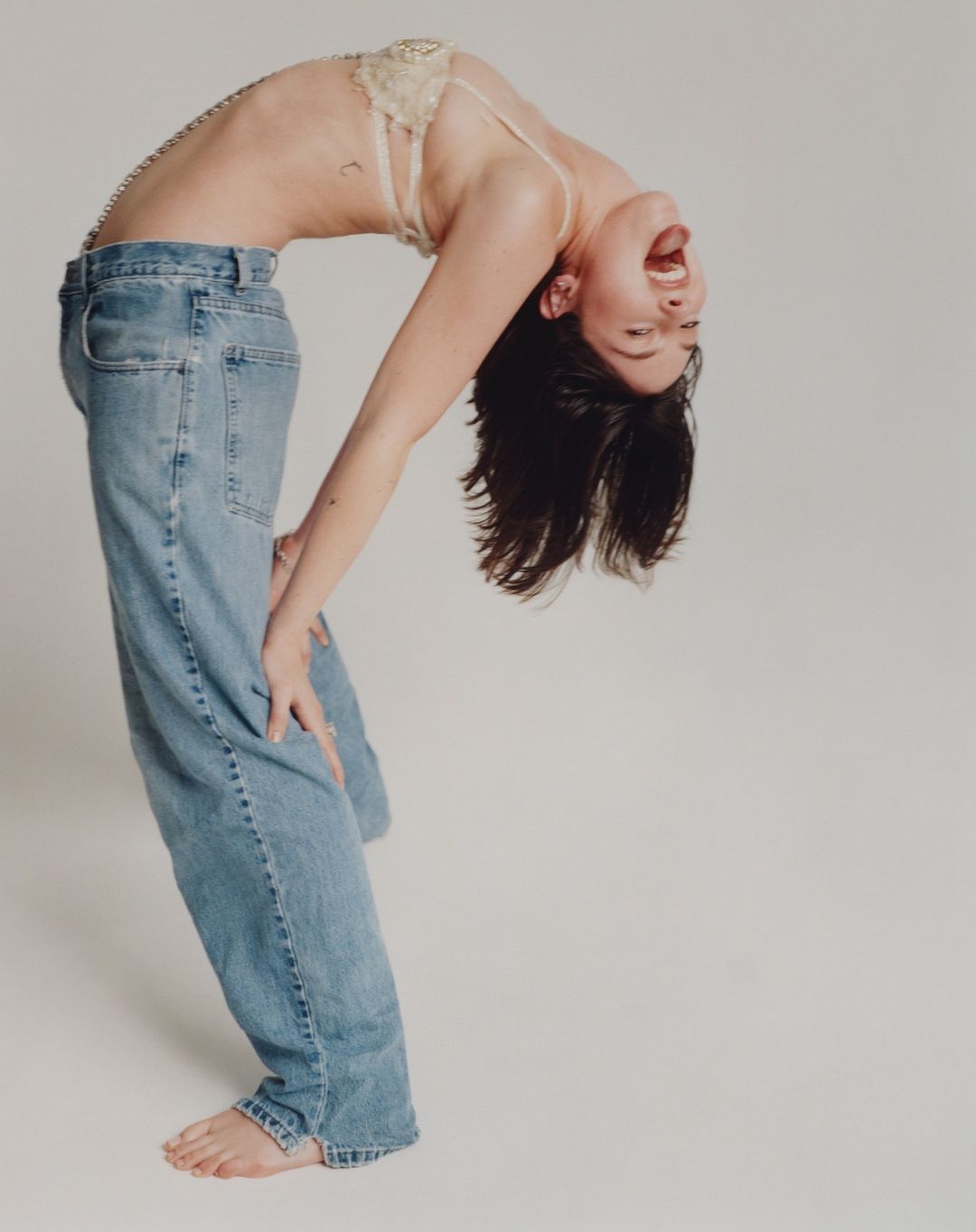 Woman bends backward in jeans and a bra in a photo studio, showcasing a dynamic pose with a minimalist background.