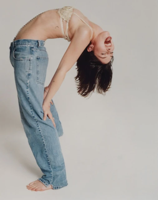 Woman bends backward in jeans and a bra in a photo studio, showcasing a dynamic pose with a minimalist background.