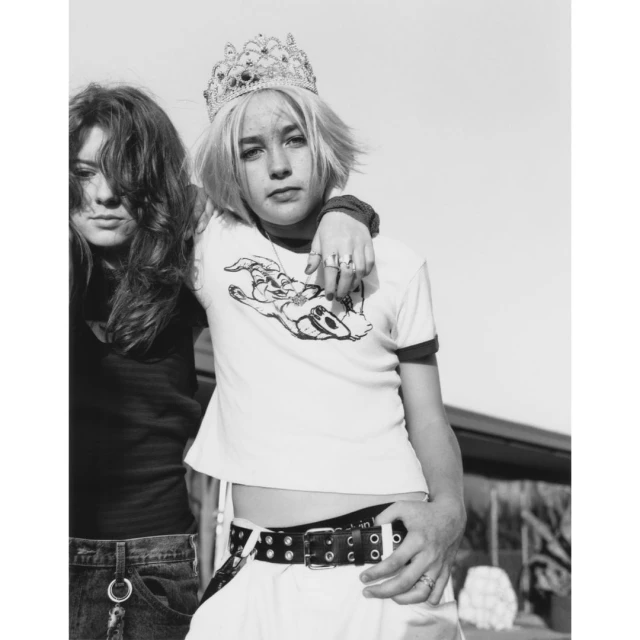Black and white portrait of two young women outside. One wears a crown and white t-shirt with graphic. The other is beside her.