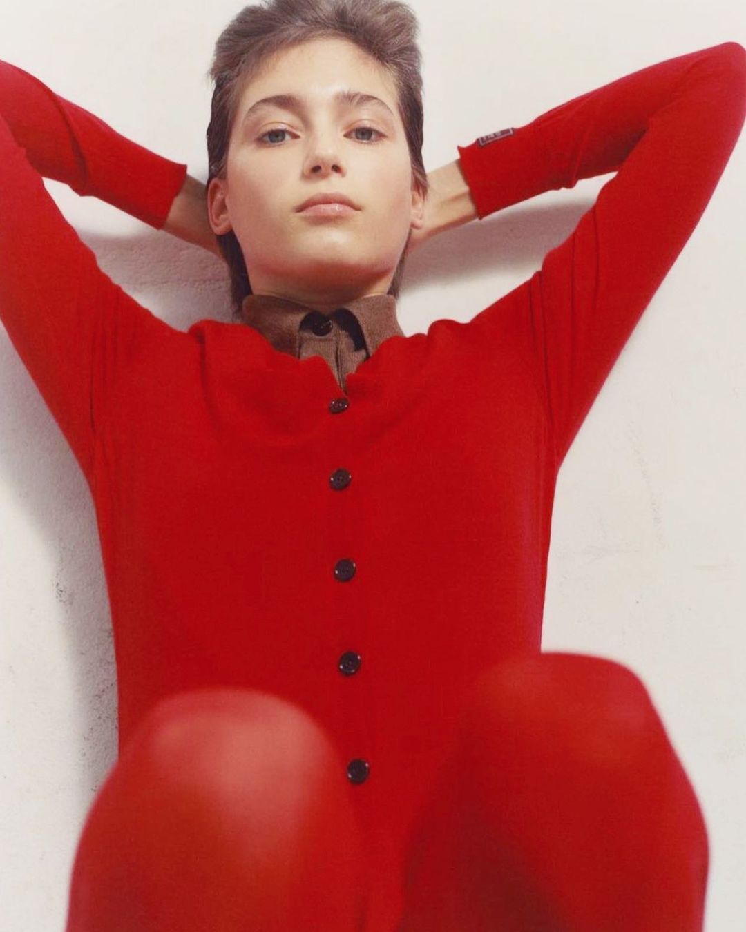 Woman in red cardigan with hands behind her head, posing against a neutral backdrop in a fashion or portrait photography setting.