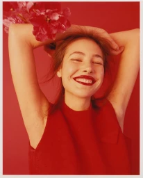 Smiling woman in a red top holding pink flowers against a solid red background photographed by Clara Balzary.