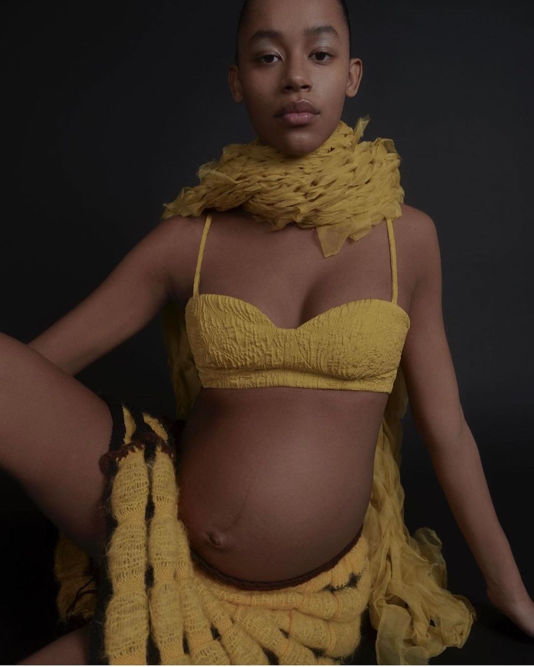 Pregnant African-American woman in a yellow knit bra top, scarf, and skirt poses against a dark backdrop in a fashion shot.