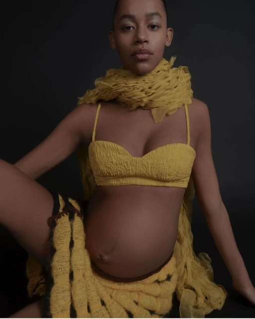 Pregnant African-American woman in a yellow knit bra top, scarf, and skirt poses against a dark backdrop in a fashion shot.