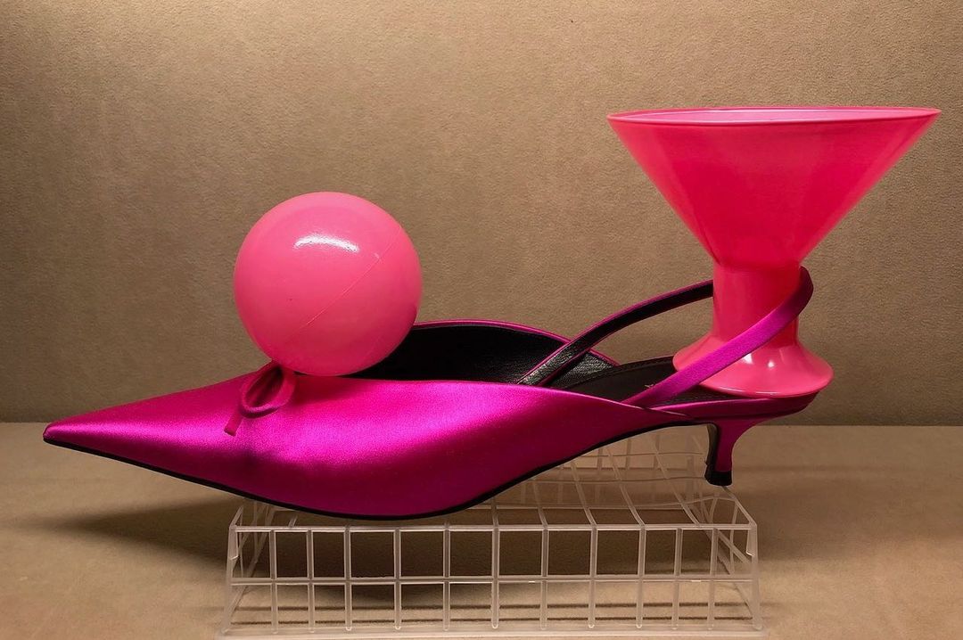 Pink satin mule shoe with a pink plastic cocktail glass and ball balancing on top, displayed on a wire grid against a tan backdrop.