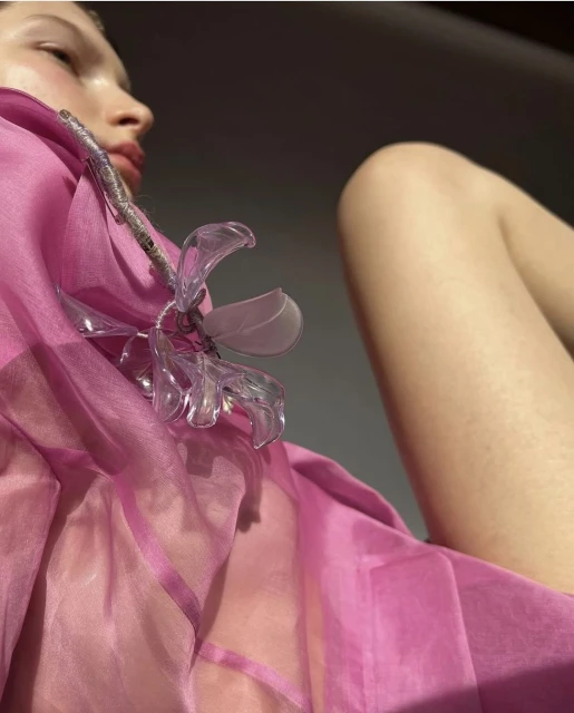 Woman poses in a pink, semi-sheer outfit, paired with a plastic flower embellishment, under a combination of bright and diffused lighting.