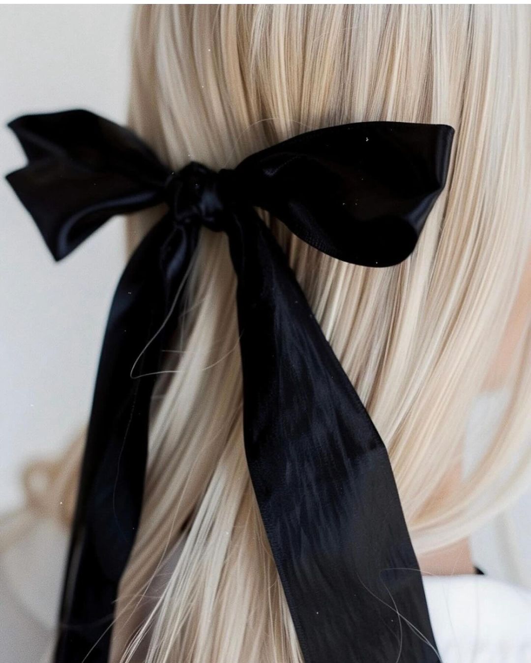 Close-up of blond hair styled with a large black ribbon bow, showcasing an elegant and simple hairstyle against a neutral background.