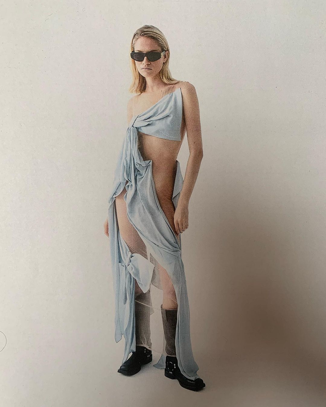 A slender, fair-skinned woman stands in a photo studio wearing a light-blue draped dress, black sunglasses, and black platform boots.
